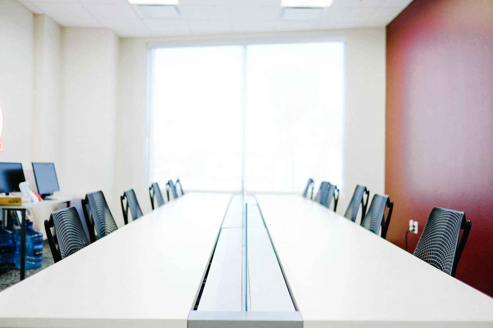 Salle de réunion vide et lumineuse, longue table blanche, chaises noires et mur rouge