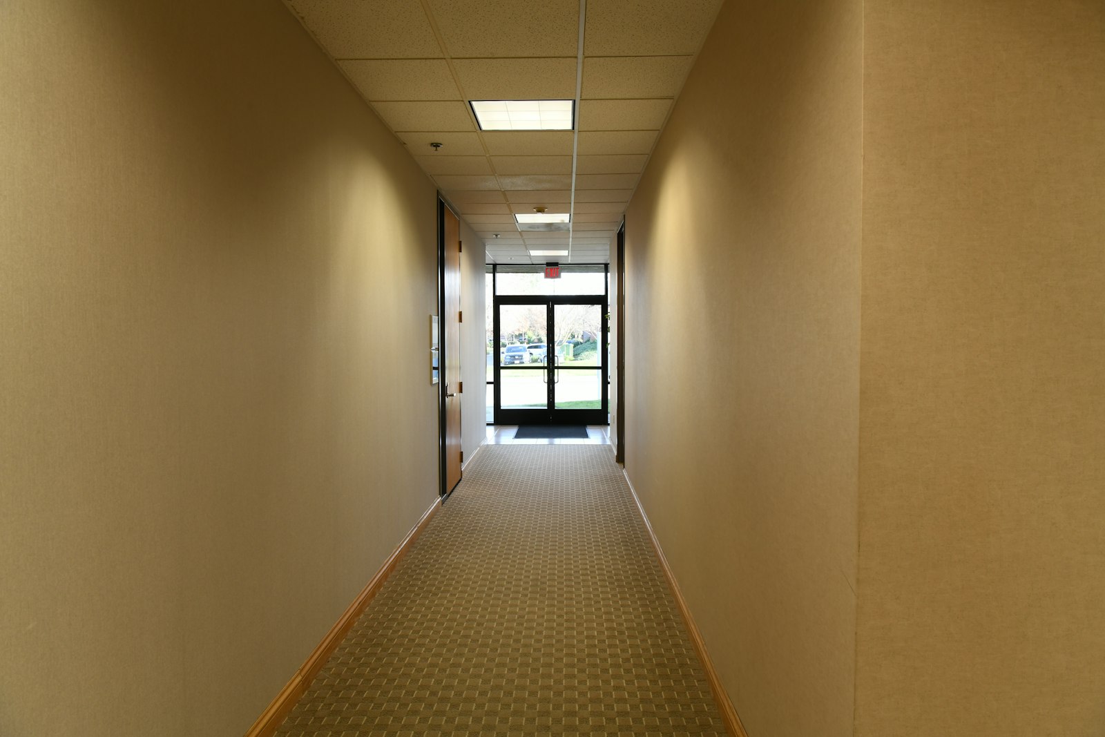 Long couloir de bureaux aux murs beiges, une porte vitrée au fond ouverte sur l'extérieur